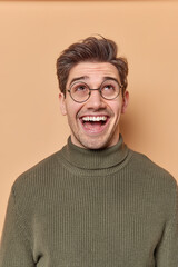 Obraz premium Vertical shot of handsome cheerful European man focused above on ceiling laughs positively sees something funny overhead dressed in casual jumper isolated over brown background. Wow what great offer