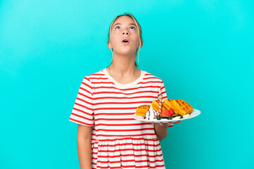 Little caucasian girl holding waffles isolated on blue background looking up and with surprised expression