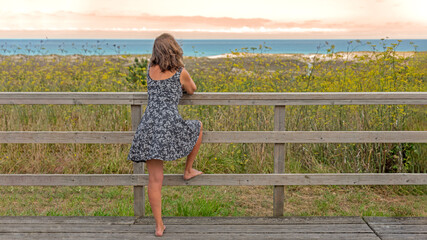 Barefoot blonde Caucasian woman on wooden walkway in short printed dress looking out to sea