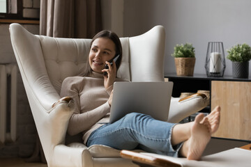 Positive busy young entrepreneur woman resting in armchair, working from home using laptop computer, talking on cellphone. Freelance worker, remote employee giving consultation on phone. Multitasking