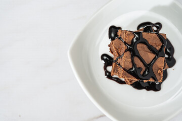 pile of chocolate brownies with strands of chocolate sauce on a white plate on a marble table. Copy space.