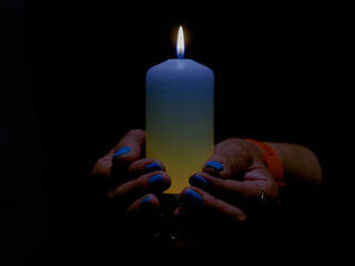 Płonąca świeca trzymana w kobiecych dłoniach. Świeca ma odcień niebieski i żółty. Na takie same kolory pomalowane są paznokcie. Jest to symbol solidarności z ofiarami wojny w Ukrainie, - obrazy, fototapety, plakaty