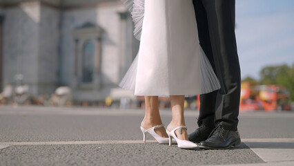 Legs of stylish bride and groom on the background of city summer street with driving cars. Action. Man in black suit and a woman in white dress.