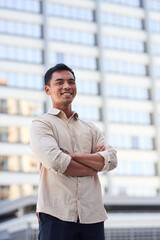 A young attractive Asian man stands in front of city buildings with arms crossed