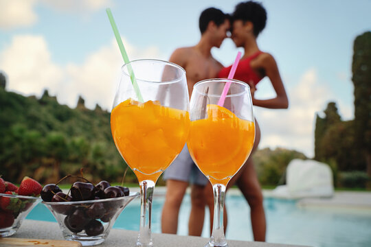 two glasses with iced orange fruit, some cherries and strawberries against a blurred multiethnic couple with African American girlfriend and caucasian boyfriend touching foreheads by the swimming pool - Powered by Adobe