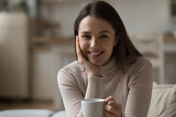 Happy pretty girl drinking morning tea, cocoa, chocolate, holding cup of hot beverage, enjoying home leisure, looking at camera, smiling, relaxing. Head shot portrait