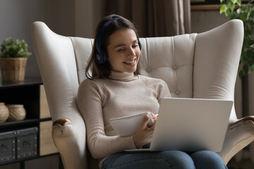 Positive happy gen Z student girl wearing headphones, talking to teacher, tutor in video call, watching learning webinar, class, lesson on laptop, attending college conference, writing notes