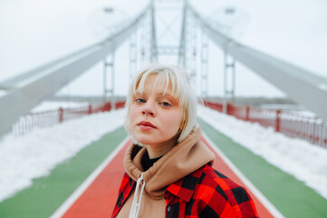 Obraz premium Portrait of a stylish hipster girl with blond hair on a winter walk standing on the street on a background of snowy views.