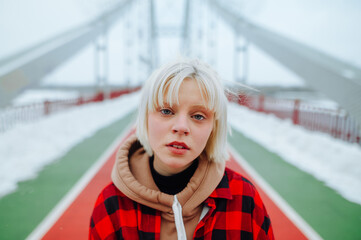 A close-up portrait of a girl in a blonde-haired shirt standing on a snow-covered bridge in winter and posing for the camera with a serious face.