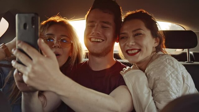 Beautiful young girls taking selfie with their friend on phone or smartphone in back seat of moving car while roadtrip. Cheerful friends having good time together. Concept of joy and lifestyle