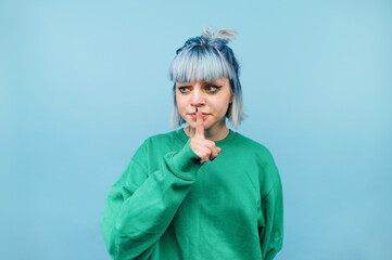 Cute teen girl with blue hair stands on a blue background, looks away and shows a gesture of silence.