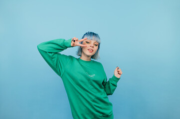 Beautiful girl in a green sweatshirt and with colored hair poses for the camera on a blue background, shows a gesture of peace and laughs