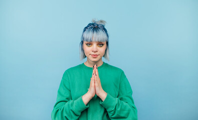 Beautiful hipster girl with blue hair in a green sweater stands on a blue background and prays with...