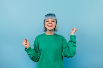 Joyful hipster girl with blue hair stands on a blue background with a happy face raises her hands up and smiles, wears a green sweatshirt
