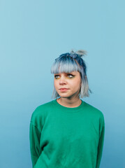 Pensive hipster girl with colored hair stands on a blue background with a serious face looking up. Vertical