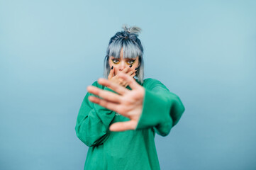 Shocked scared girl in casual clothes stands on a blue background and shows his palm to the camera gesture stop and covers his face with his hand