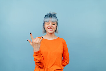 Positive girl in an orange sweatshirt stands on a blue background with closed eyes and shows a gesture of heavy metal with an expressive face.