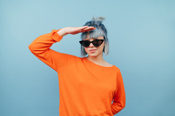 Serious hipster girl in sunglasses stands on a blue background and looks to the side hiding his hand from the sun.