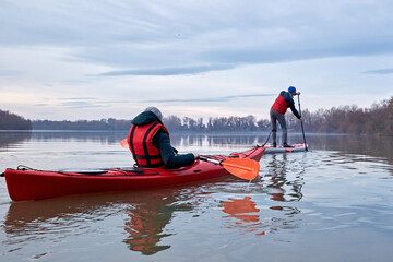 Woman in the red kayak and man paddle on stand up paddle boarding (SUP) rowing on the Danube river on cloudy calm winter day