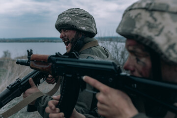 Two Ukrainian soldiers scream in rage and shoot from kalashnikov assault rifles to russian enemy.