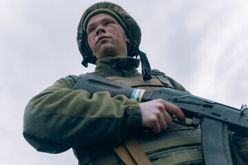 Portrait of Ukrainian soldier in helmet with kalashnikov assault rifle against sky background.