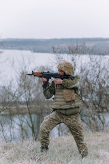 Ukrainian soldier aims from kalashnikov assault rifle to russian enemy against background of river and sky.