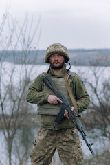 Ukrainian soldier stands with kalashnikov assault rifle in his hands against background of sky and...