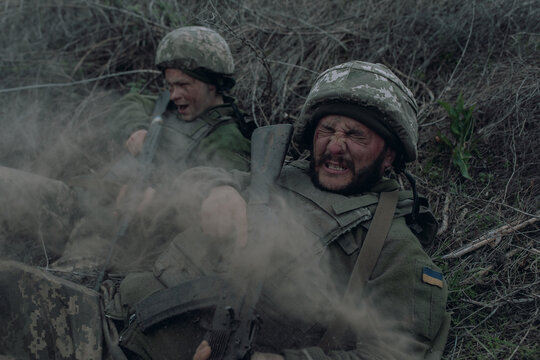 Two Ukrainian soldiers lie with kalashnikov assault rifles on dry grass during artillery bombardment.