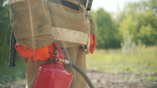 Close up side view of firefighter with a red fire extinguisher in a body safe suit uniform for protection from fire operation. Clip. Male fireman standing in the green field.
