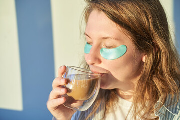 Young woman drink coffee at home. Morning routine. Eye patch for skin health. Mug with beverage. Female person and transparent cup. Hydrogel cosmetic treatment