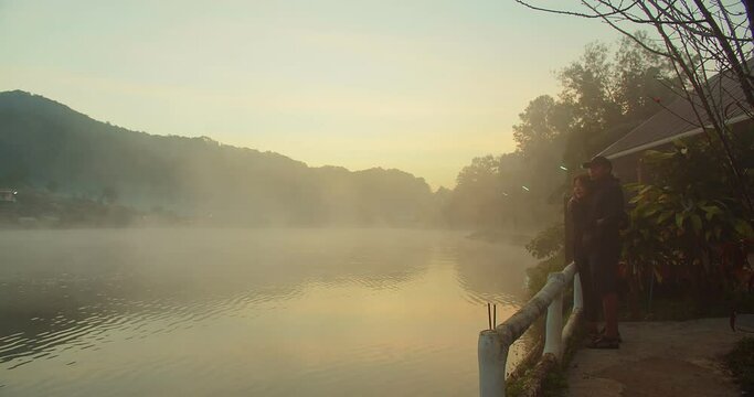 happy young Asian couple tourists travelers standing enjoy vacation holiday trip against sunrise in the morning at lakeside ban rak thai village, Mae Hong son Thailand, travel together adventure