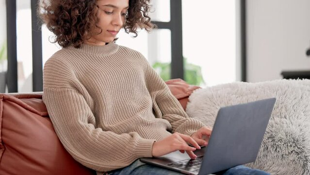 young mixed race woman with curly hair typing on laptop wearing in cozy casual sweater sitting on the coach at home