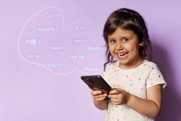 A little joyful brunette child stands on a purple background with a phone and learns different...