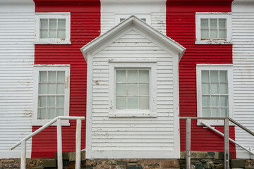 The facade of a red and white striped lighthouse outcrop building. The center is a porch with a small glass window containing nine panes of glass. There are two windows on both sides of the porch. 