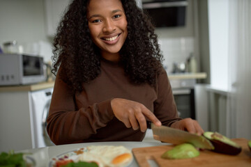Smiling black woman cutting vegetables sitting at table in front of plate with fried eggs, wooden board, slicing peeled avocado against modern kitchen interior, wearing brown sweater