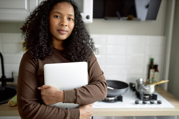 Adorable young dark-skinned girl holding silver portable personal computer, standing against cooker at kitchen, going to sit at table to have breakfast and work from home in early morning