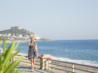 Fototapeta premium A seascape against a backdrop of mountains with a beautiful sky in sunny Turkey. A blonde girl walks along the seafront in a short dress. A young woman looks out toward the sea, watching the waves.