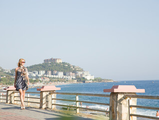 A seascape against a backdrop of mountains with a beautiful sky in sunny Turkey. A blonde girl walks along the seafront in a short dress. A young woman looks out toward the sea, watching the waves.