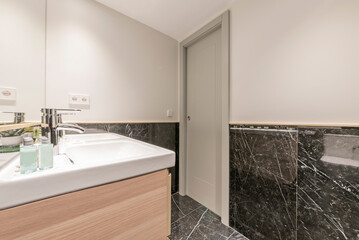 Bathroom with wooden furniture, porcelain sink and frameless mirror embedded in the wall and veined black marble on the walls and floor