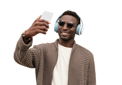 Portrait of happy smiling young african man taking selfie by smartphone while listening to music in headphones isolated on white background