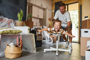 Portrait of carefree father playing with son riding on chair in hall, happy family moving into new...