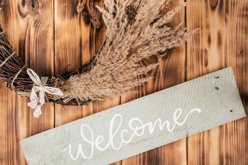 Dry herbal, cereal tightly woven wreath with string and nameplate welcome. Rustic brown wooden and planking door on background. Celebration and preparation, warm greeting of guests. Cosy composition