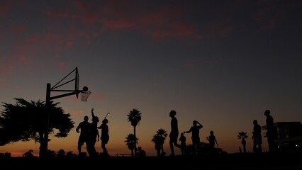 Naklejka na ściany i meble People playing basket ball game, silhouettes of players on basketball court outdoor, sunset ocean beach, California coast, Mission beach, USA. Black hoop, net and backboard on streetball sport field.