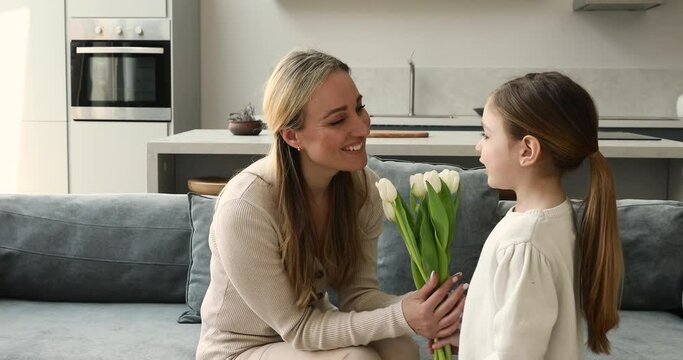 Grateful excited single mother sit on couch receive greetings with bunch of tender spring tulips from beloved little daughter. Adorable preschool girl congratulate mommy with March 8 give kiss flowers