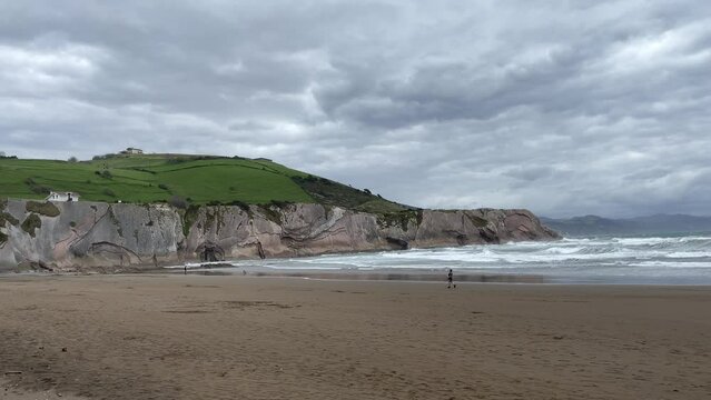 Lonely person walking on Itzurun beach by cliff coast and rough sea on cloudy day in Zumaia. Alone in nature, freedom concepts