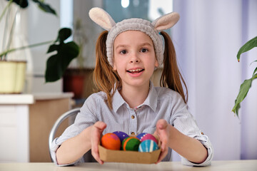 A positive playful girl in bunny ears holds colorful eggs in her hands in the home kitchen. Happy Easter. Easter background