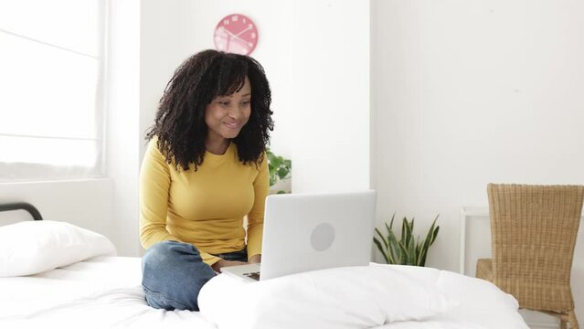 Happy young african american woman relaxing on bed having a video call. Cheerful brazilian female having a conversation through videoconference app on laptop computer - High quality 4k footage