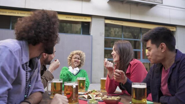 Happy young modern friends enjoying time together eating tapas and drinking beer in outdoors bar celebrating holiday trip