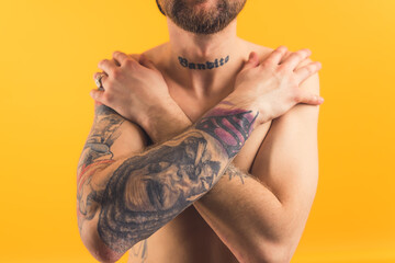 Tattooed shirtless caucasian adult man crosses arms in front of his chest covering his body over yellow background. Body shaming concept. High quality photo