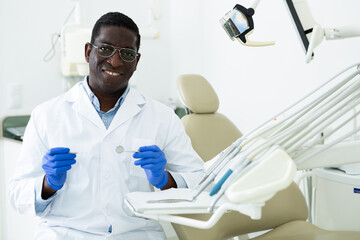 Portrait of successful smiling professional african american dentist standing in white coat in...